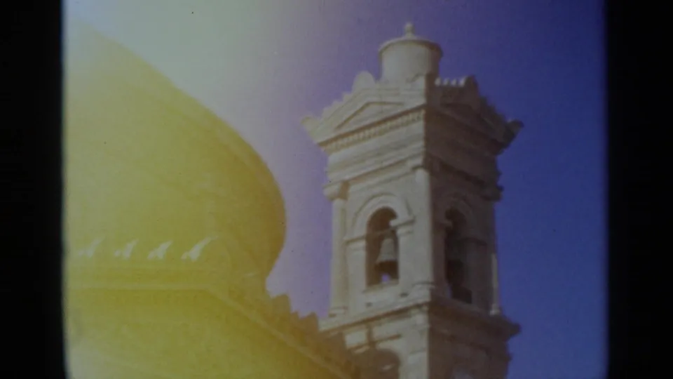 1960s 8mm travel view of an ornate bell tower rising beside a large domed church roof, with yellow exposure flare across the frame.