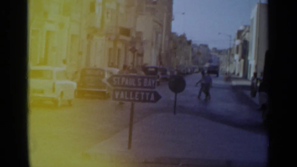1960s 8mm street view with pedestrians, parked cars, masonry buildings and a directional sign reading St Paul's Bay and Valletta.