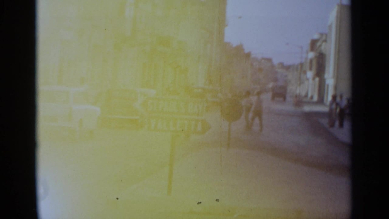 1960s 8mm street view with pedestrians near cars and a directional sign, with strong yellow color cast and soft exposure.