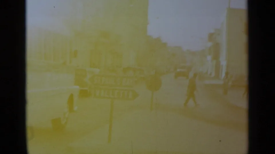 Faded 1960s-style 8mm frame showing a pedestrian crossing near a directional sign, parked cars, and sunlit buildings.