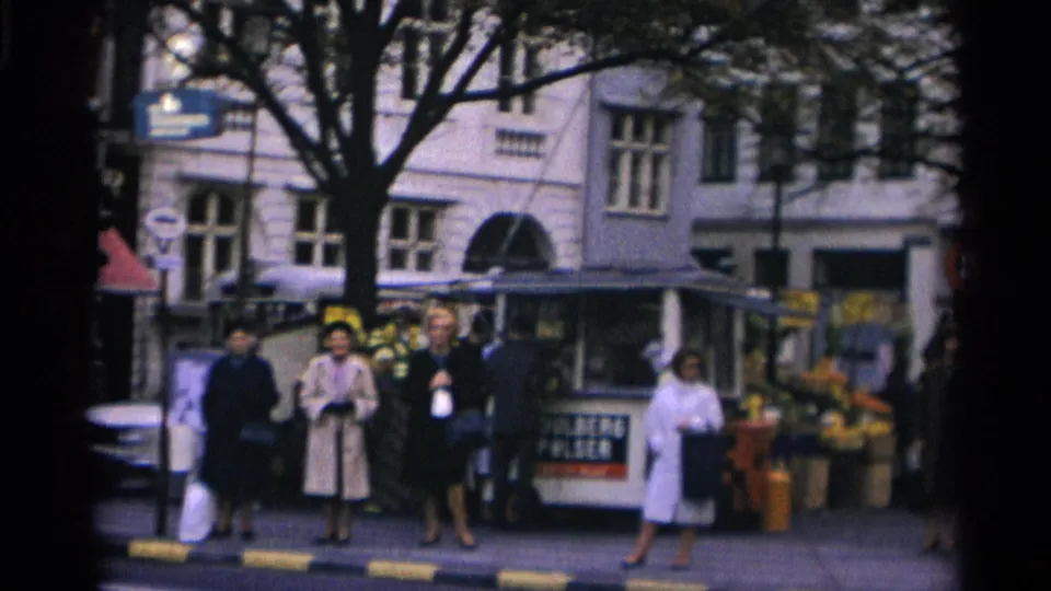 1966 Copenhagen street corner with women in coats standing near a small kiosk, flower display, pedestrians, trees, and storefront windows.