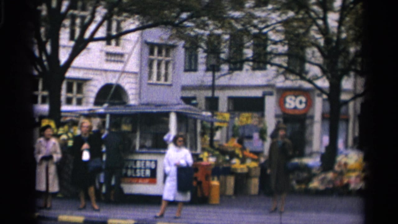 Copenhagen street scene with women in period coats, a flower stall, kiosk, pedestrians, trees, and a visible round storefront sign.