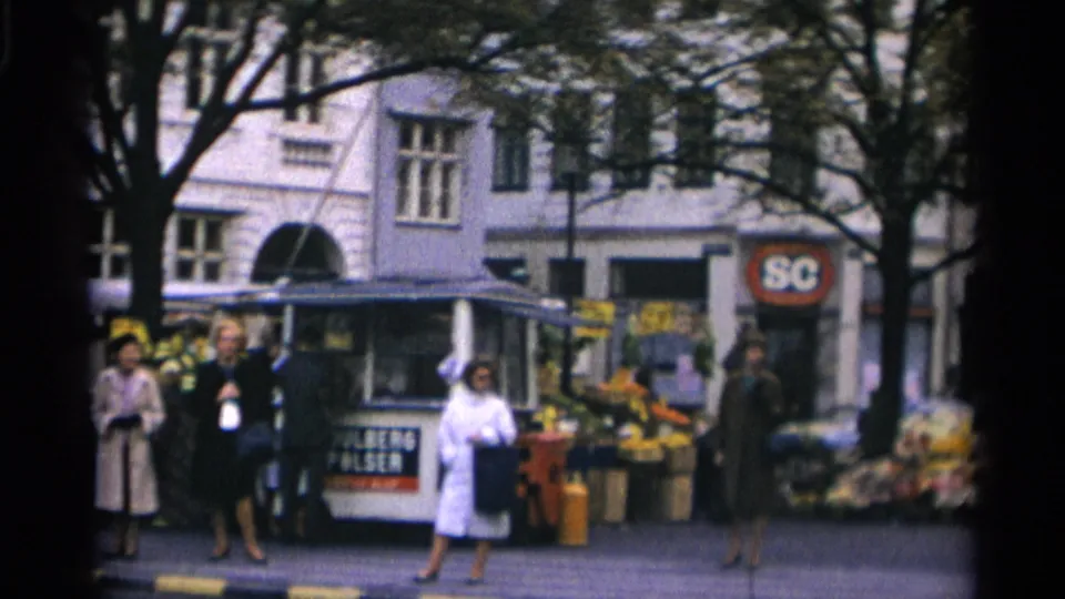 Copenhagen street scene with women in period coats, a flower stall, kiosk, pedestrians, trees, and a visible round storefront sign.