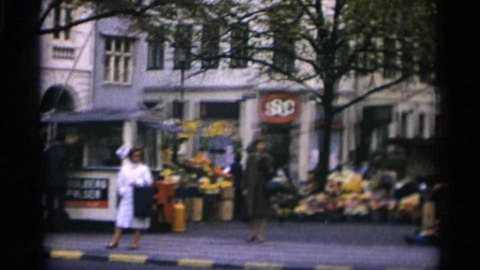 A woman in a light coat stands near a Copenhagen kiosk, with flowers, produce crates, pedestrians, and a round storefront sign behind her.