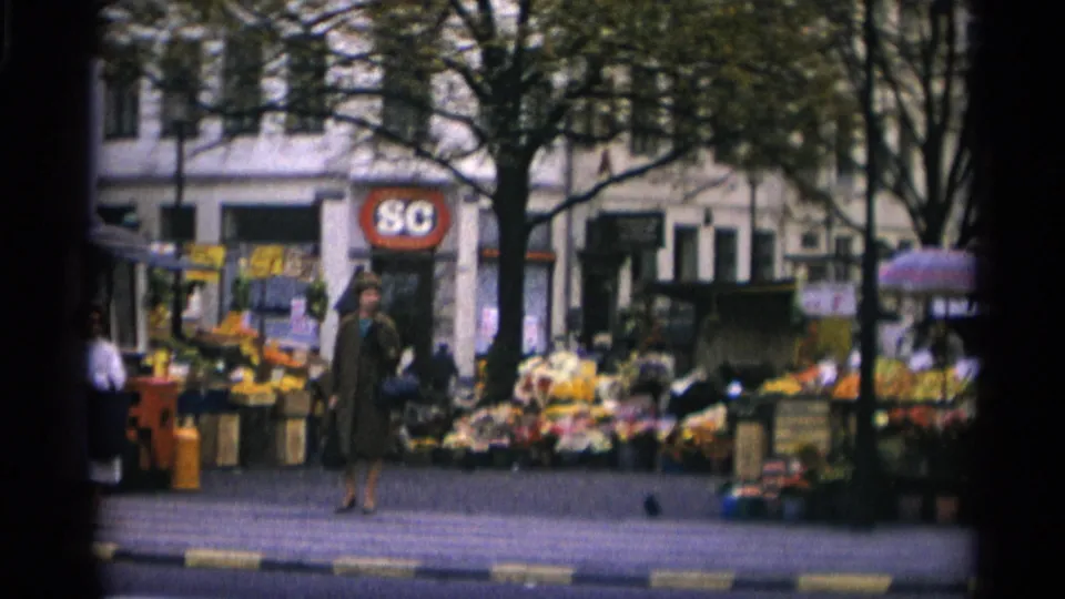 1966 Copenhagen street view with a woman walking near flower displays, a kiosk, pedestrians, trees, storefronts, and curb markings.