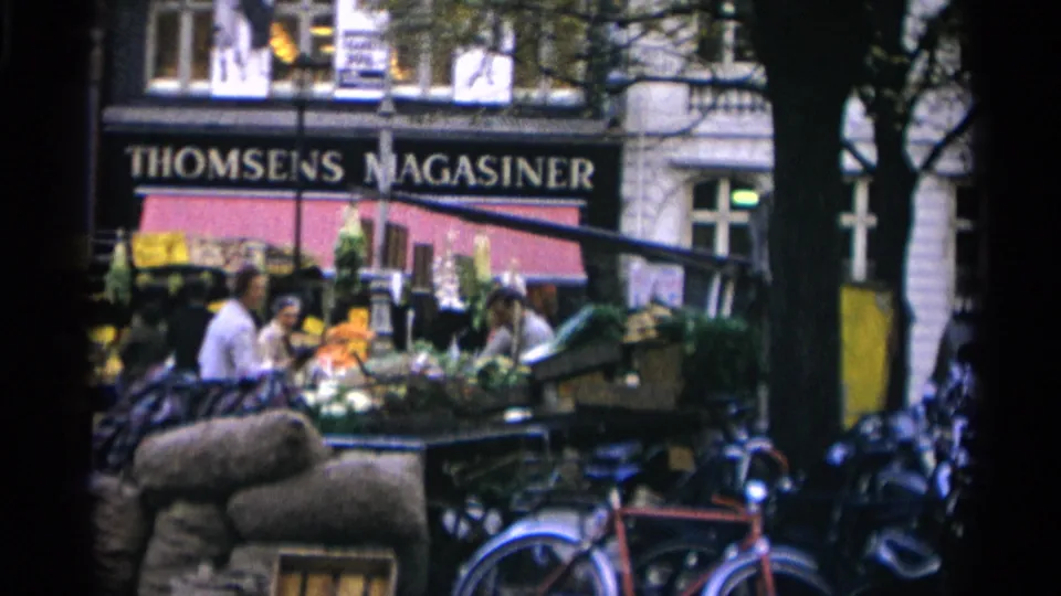 1966 Copenhagen market frame with produce and flowers, several shoppers, large sacks, parked bicycles, and the THOMSENS MAGASINER sign.