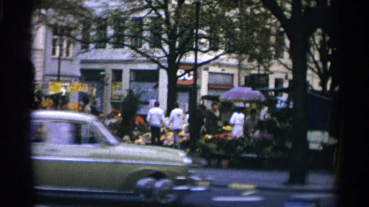 1966 Copenhagen street view with a pale sedan crossing the foreground and pedestrians gathered near outdoor stalls behind trees.