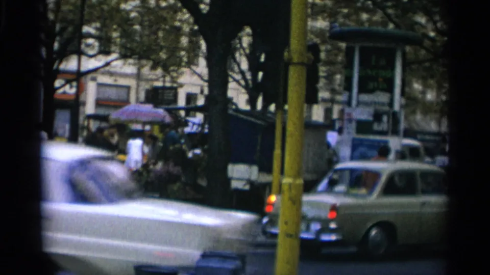 Copenhagen street traffic in 1966, with a blurred car crossing the frame near a yellow pole and pedestrians by a bus stop area.
