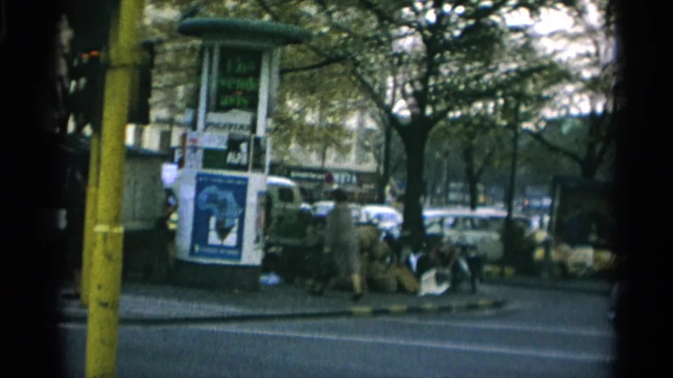 1966 Copenhagen street corner showing a yellow pole, poster kiosk, parked cars, and people beside a tree-lined roadway.