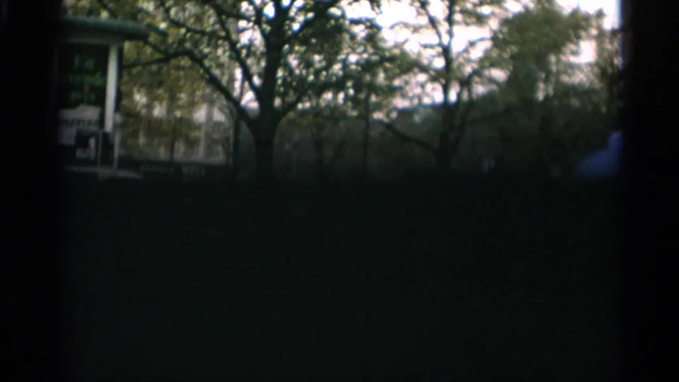 Dim final 8mm frame of the Copenhagen streetscape, with trees, buildings, a poster sign, and dark foreground framing.