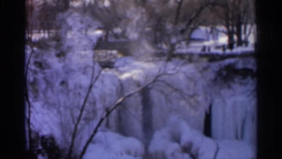 8mm winter view of Minnehaha Falls area, with icy falling water, snow-covered banks, bare branches, and a distant structure.