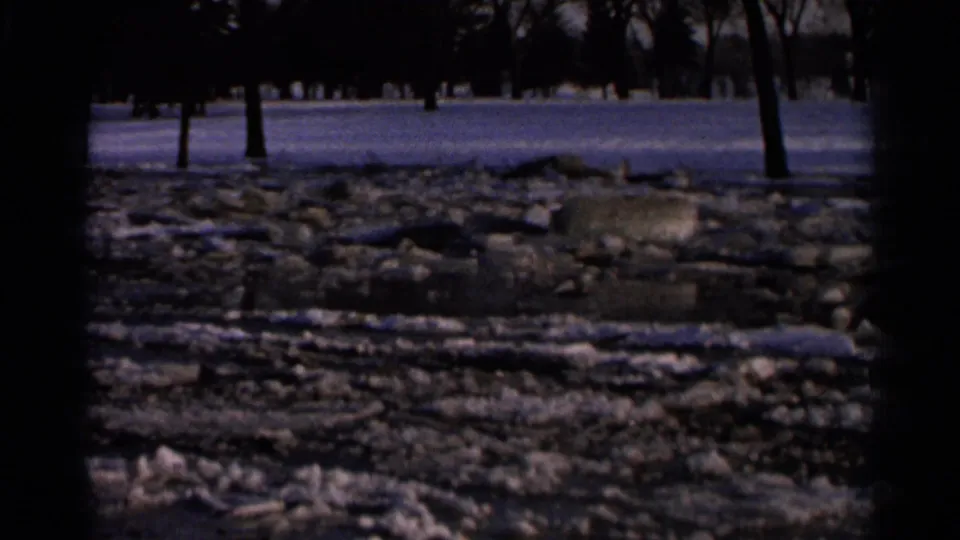 1966 Hamel, Minnesota 8mm frame of a half-frozen pond with broken ice, exposed stones, snow patches, and bare trees beyond.