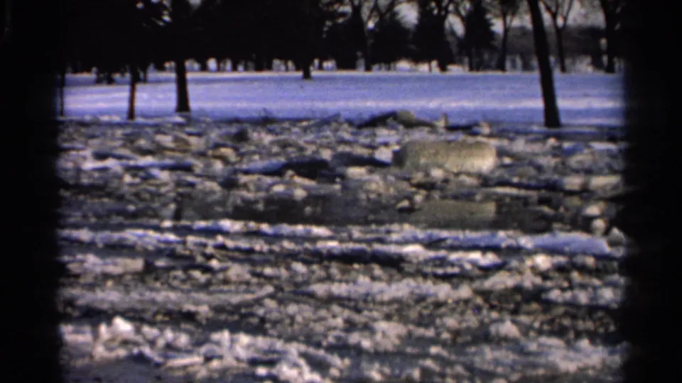 1966 Hamel, Minnesota 8mm frame showing a wintry pond surface with broken ice, snow-covered ground, rocks, and leafless trees.