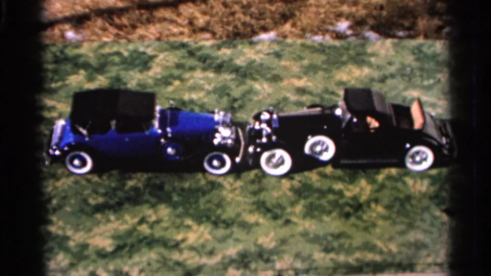 The blue and black vintage cars remain pressed together on a grassy field, with front-end contact visible in the 8mm home-movie frame.