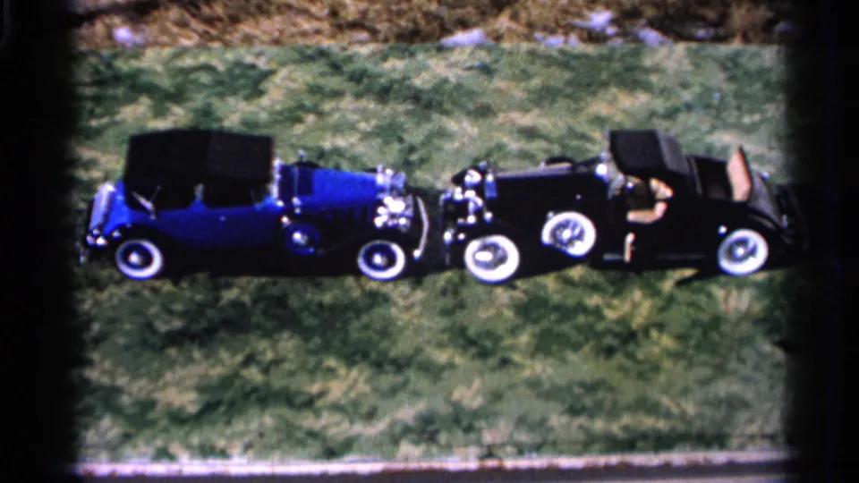 A blue convertible faces a black roadster on grass, with soft 8mm color and partial people visible in the cars.