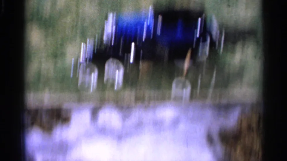 The blue toy car appears blurred by quick handheld movement as it crosses grass in the outdoor home-movie scene.