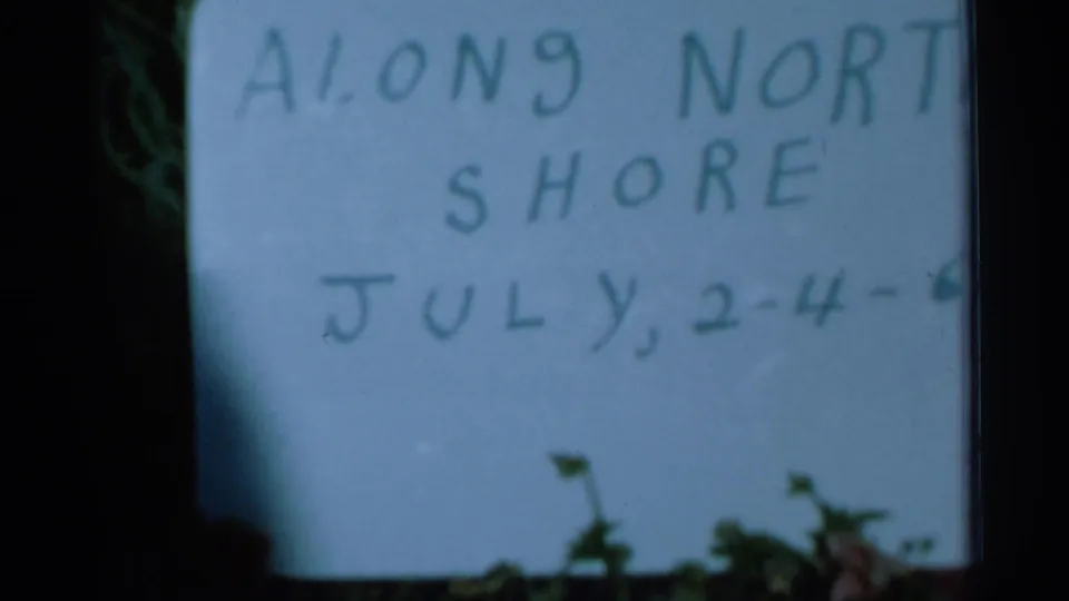 1966 8mm frame holding on the handwritten North Shore sign, with July date text visible and dark film edges around it.