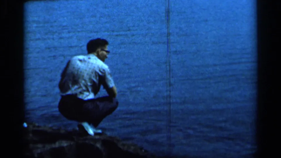 1966 Minnesota home-movie frame showing an adult man in glasses crouching at the shoreline beside dark blue water.