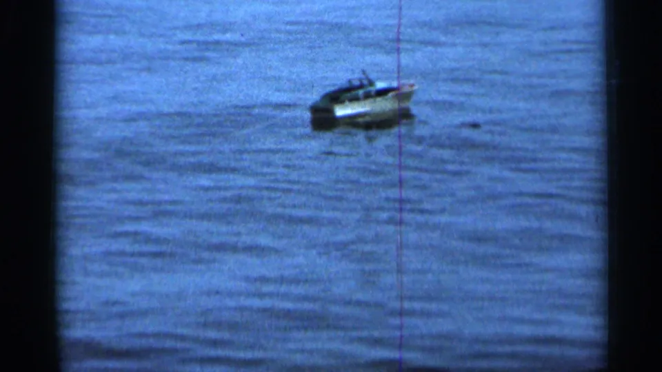 1966 8mm home-movie frame showing a small boat floating on rippled water, viewed from a distant shoreline position.