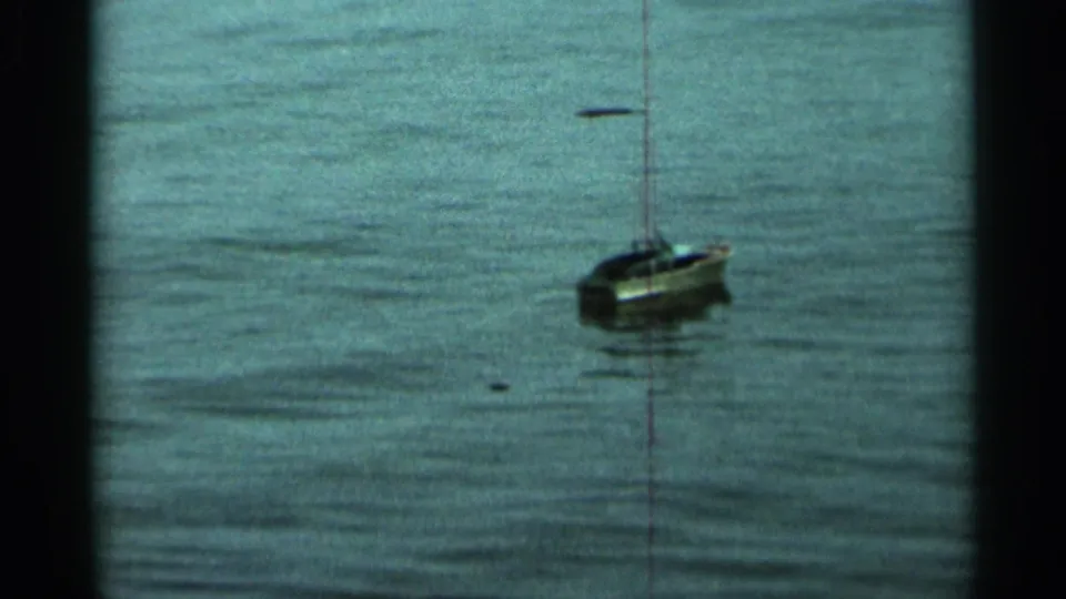The small sailboat continues drifting on the lake, with faded color and visible frame edges from the 8mm home-movie scan.