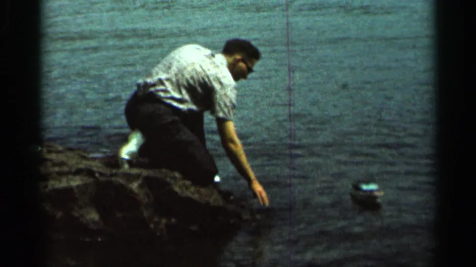 An adult man in a light shirt kneels on a rocky lakeshore near the toy boat, reaching toward the water in 1966 home-movie footage.