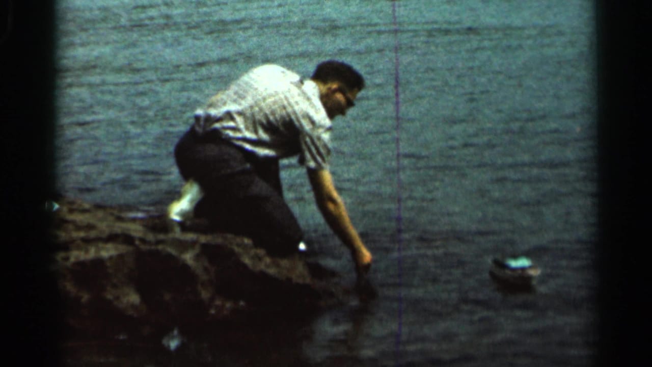 Man kneels on a rocky lakeshore while placing a small toy boat into the water.