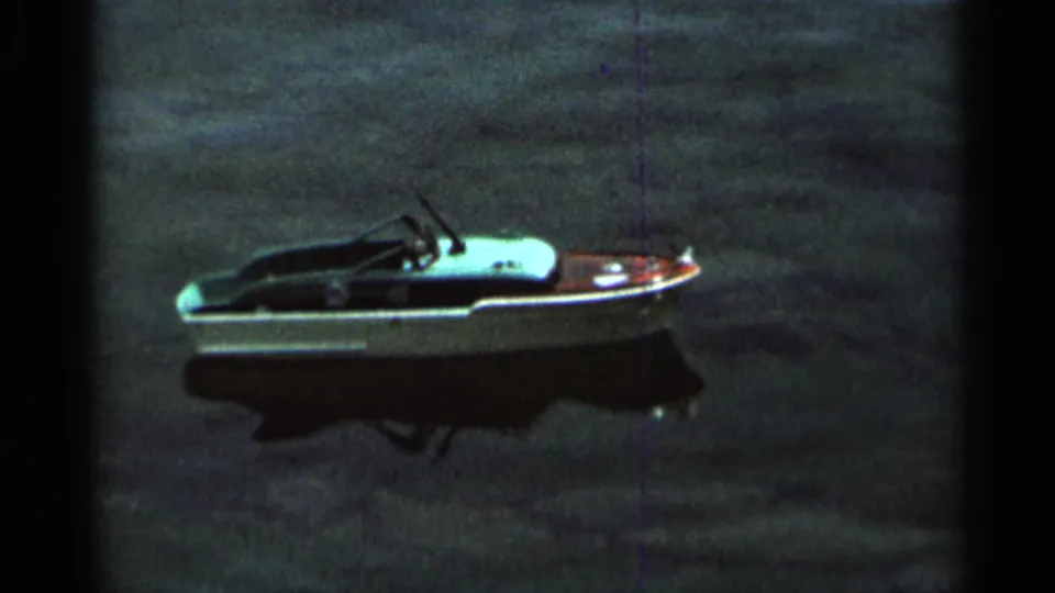 A small white-hulled toy boat drifts on lake water in 1966 Hamel, Minnesota footage, with a faint reflection on the surface.