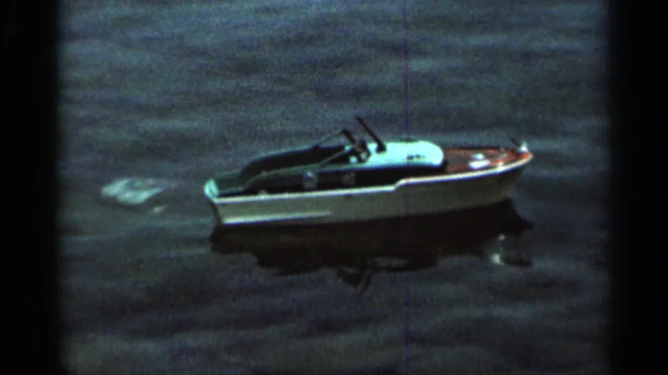 A white-hulled toy boat rests or moves slowly on the lake, with pale surface disturbance visible behind it in 1966 Hamel footage.