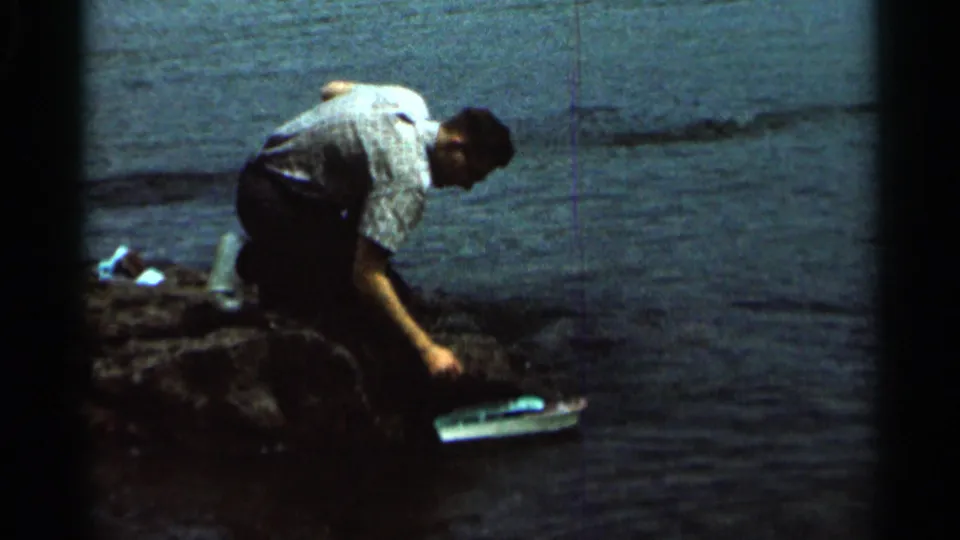 In 1966 Hamel, Minnesota home-movie footage, a man crouches at the shoreline beside rippled water and reaches toward a small toy boat.