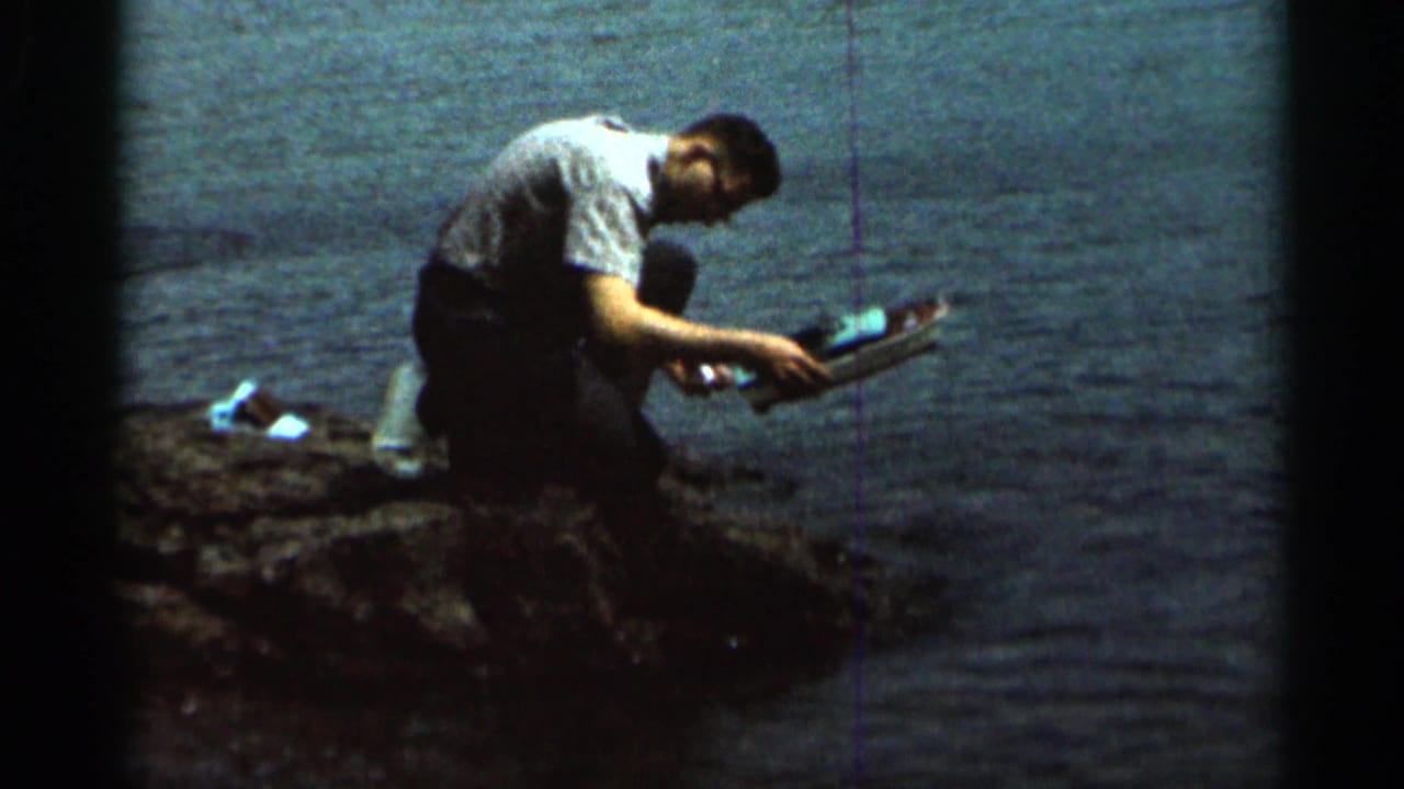 Man kneeling by water while holding a small toy boat above the shoreline.