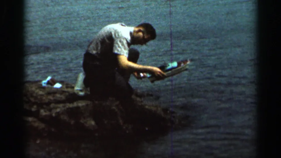 The man kneels at the water's edge in Hamel, Minnesota, holding the small toy boat as if preparing to set it into the lake or river.