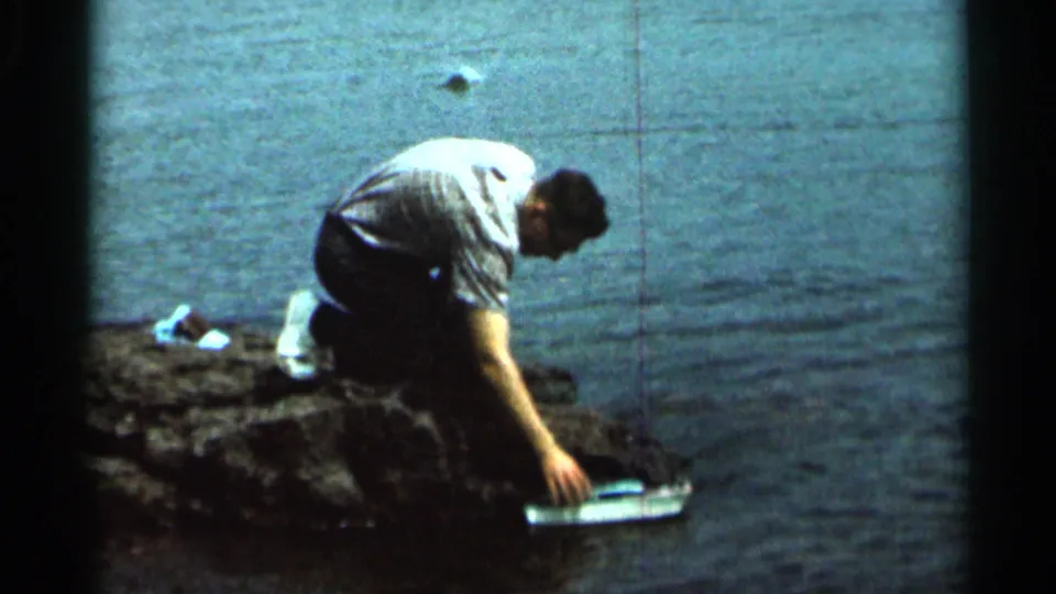 The 1966 home-movie frame shows the man leaning over the rocky shoreline and reaching to adjust the toy boat near the water.