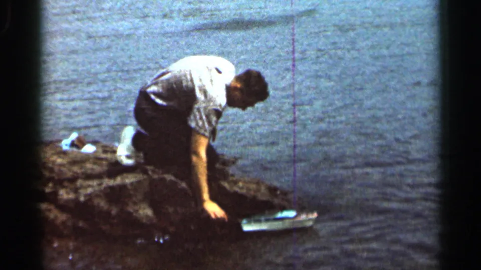 At the shoreline in Hamel, Minnesota, the man crouches with one hand near the water as the small toy boat sits close to the edge.