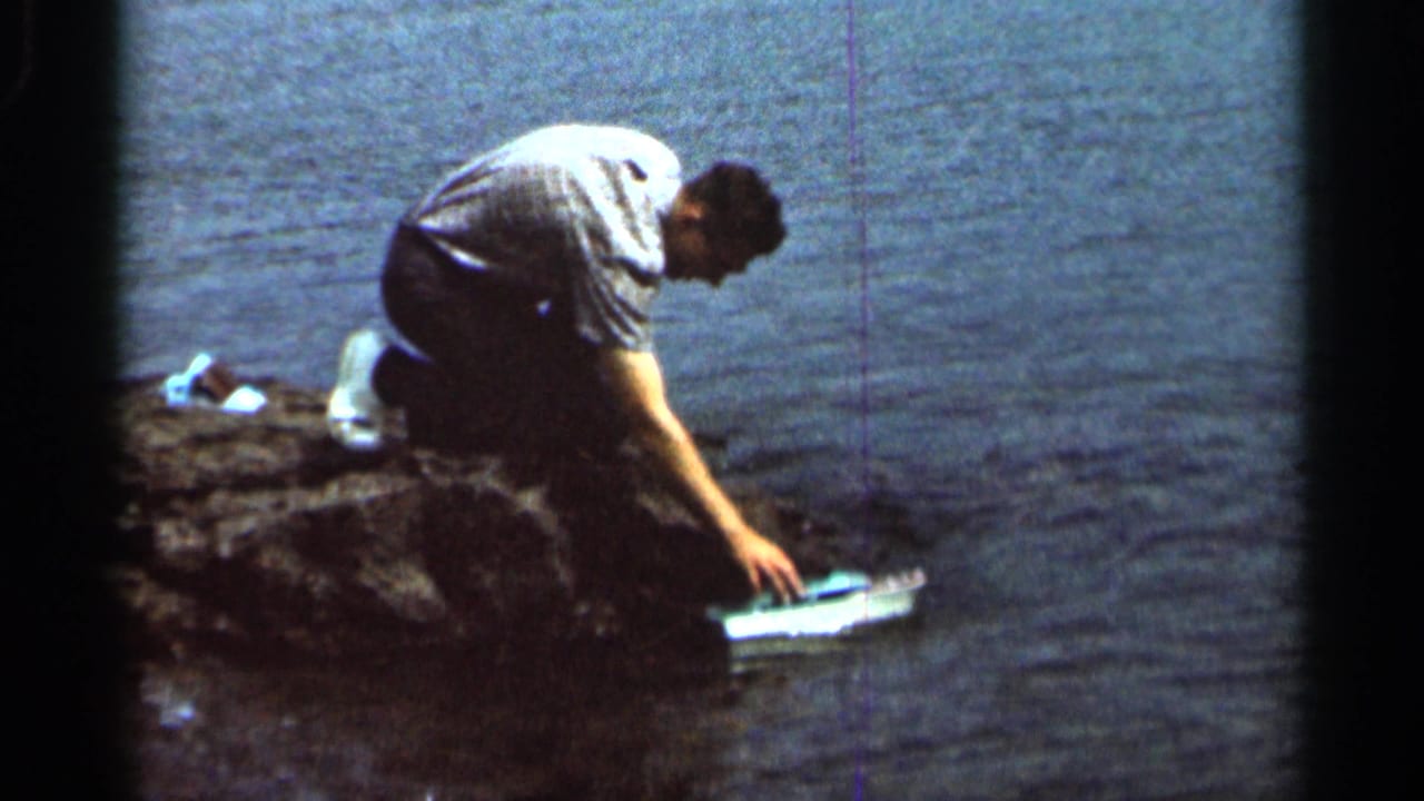 Man crouches on rocks beside a lake while reaching toward a small toy boat.