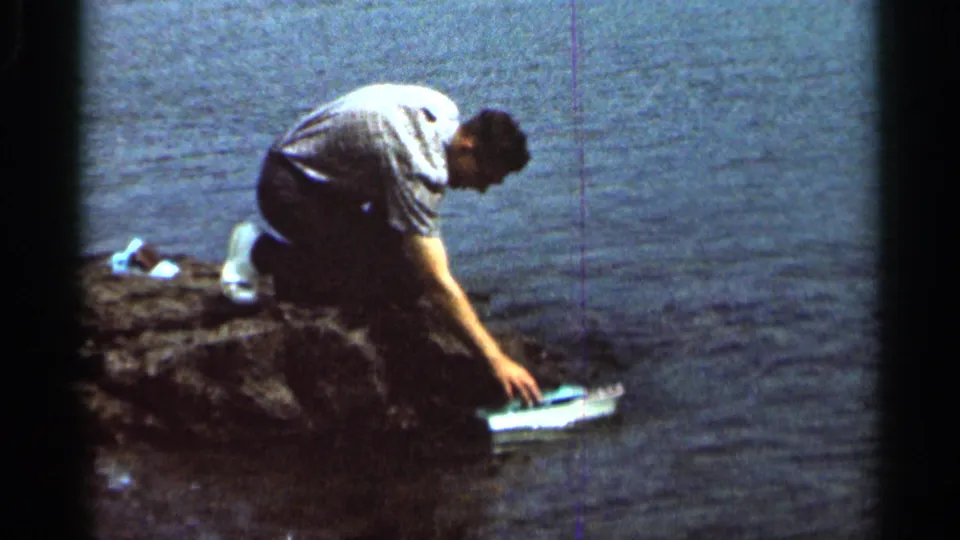In Hamel, Minnesota, 1966 home-movie footage, a man crouches at the rocky waterline and reaches toward a miniature boat.