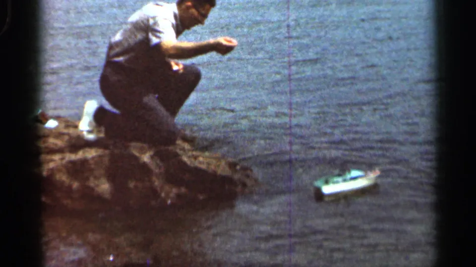 A man in a white shirt kneels on the Hamel lakeshore, holding one hand out as the small boat floats nearby.