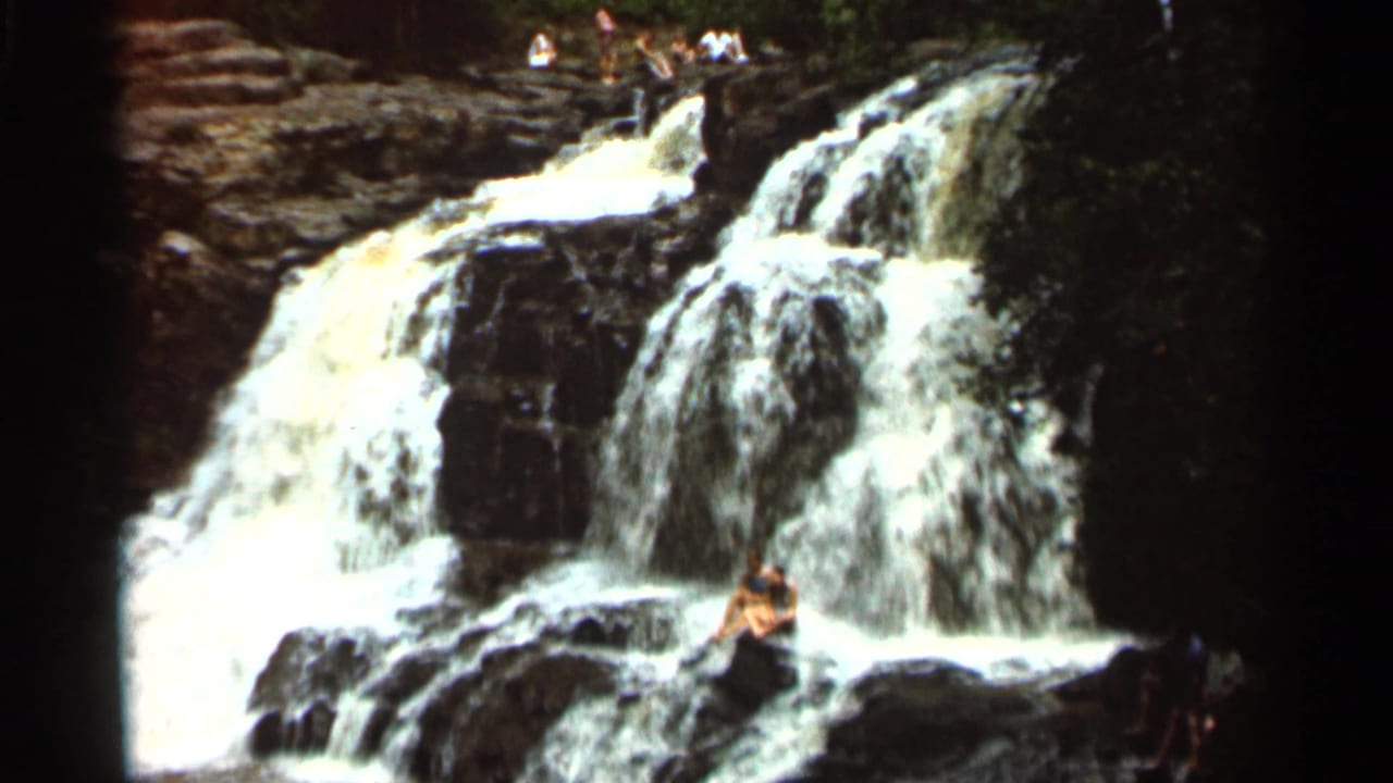 1966 8mm home-movie frame from Hamel, Minnesota, showing a waterfall, rock ledges, and a man sitting near the base.