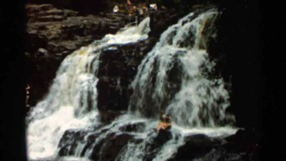 1966 Hamel, Minnesota home-movie frame showing a broad waterfall and a seated visitor on the rocks beside the rushing water.