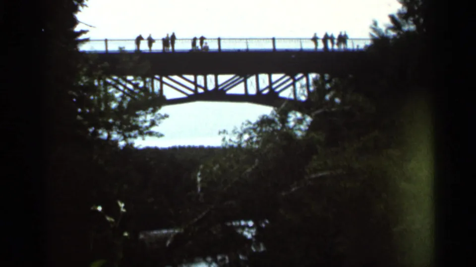 1966 Hamel, Minnesota 8mm frame showing visitors silhouetted on a pedestrian bridge above a wooded waterfall area.