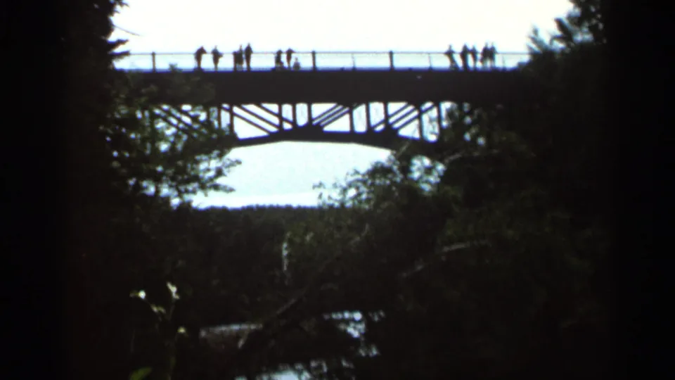 1966 Hamel, Minnesota home-movie frame with small silhouetted figures on a high arched bridge framed by dark trees.