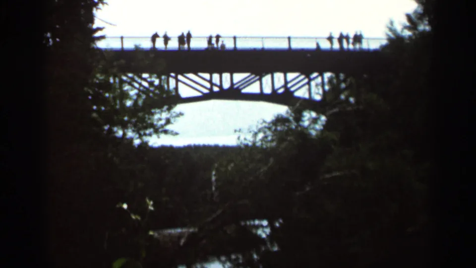 Distant 1966 home-movie view in Hamel, Minnesota showing pedestrians silhouetted on a high bridge over a wooded landscape.