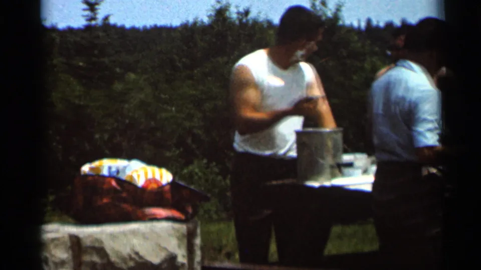 1966 Hamel, Minnesota home-movie frame showing adults in summer clothing gathered by a picnic table and cooking pot outdoors.