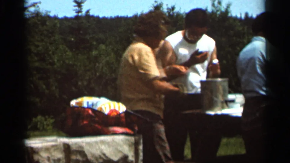 Home-movie barbecue scene from 1966 Hamel, Minnesota, with adults standing close to a picnic table and cooking pot amid trees.