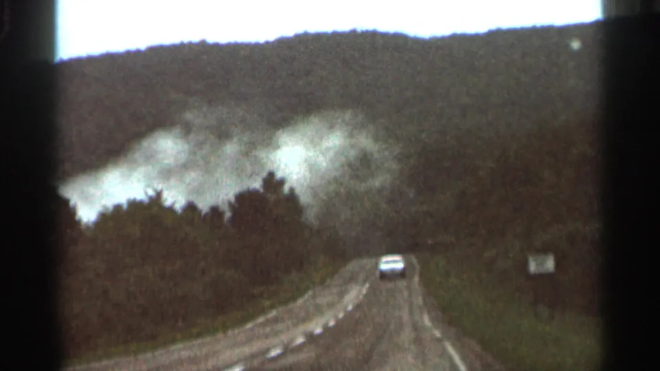 1966 Hamel, Minnesota 8mm frame showing a white car ahead on a rural road as smoke rises over wooded hills.