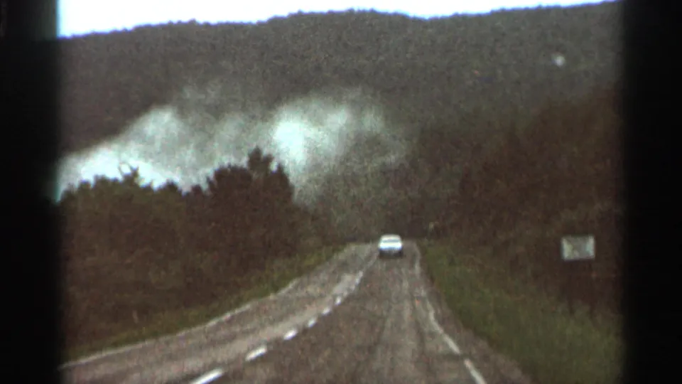 A white car continues down the rural Minnesota road with dense smoke visible above the trees in the 1966 home-movie frame.