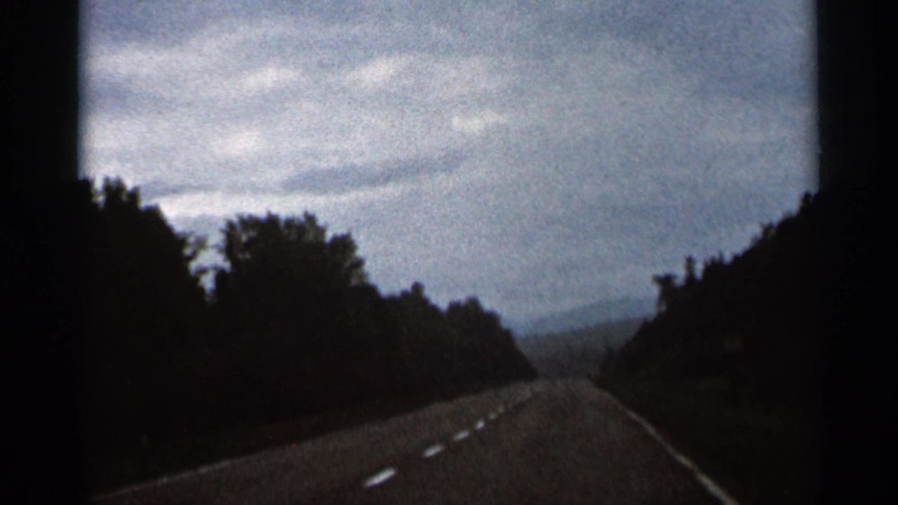 Roadway curving through dense trees with cloudy sky above.