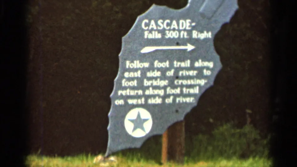 1966 8mm frame showing a Cascade Falls direction sign with trail instructions in a wooded outdoor setting.