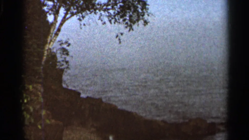 Handheld 1960s home-movie frame of rippled water, dark shoreline, and leafy branches near the Cascade Falls visit.