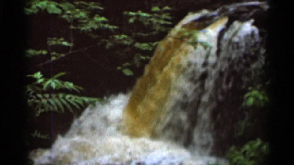 1966 8mm frame from Hamel, Minnesota showing muddy water cascading over a dark rock face amid leafy forest growth.