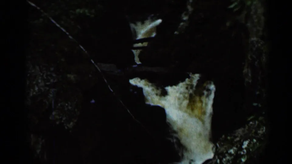 1966 8mm frame showing a narrow waterfall dropping between shadowed rocks, with branches crossing the foreground.